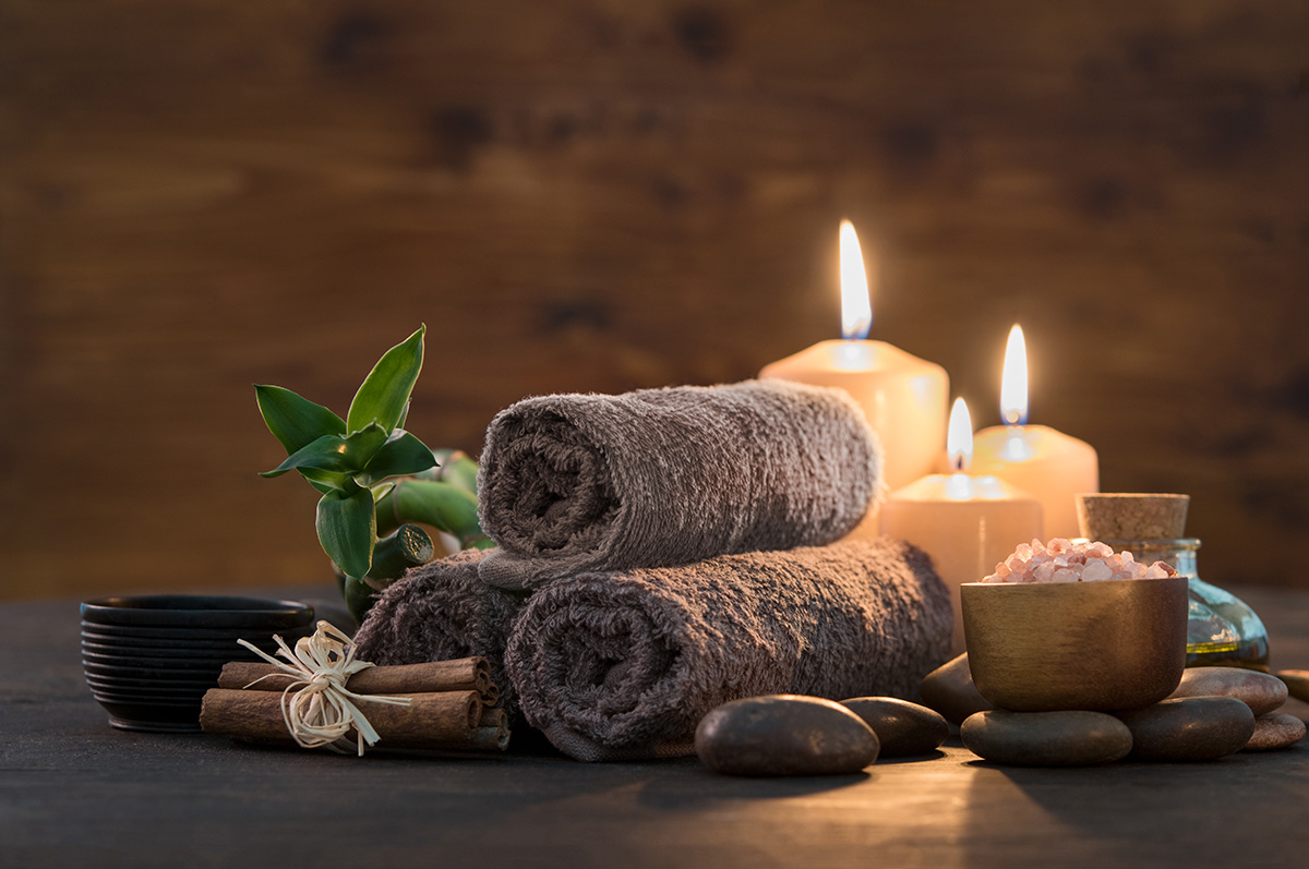 A serene spa setting with rolled dark towels, glowing candles, a green plant, smooth stones, a bowl of Himalayan salt, cinnamon sticks tied with a raffia ribbon, and massage oils creating a tranquil atmosphere.